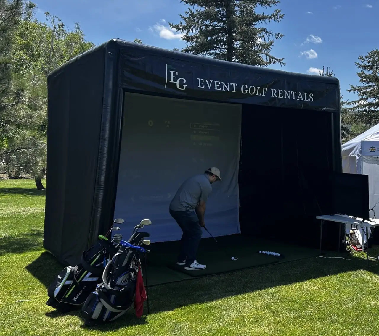 The Event Golf Rentals simulator enclosure set up on grass, with a player inside and golf bags beside it for scale